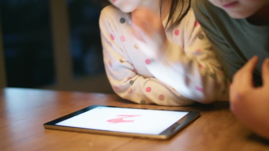 A sister and brother play a game on a tablet on a table inside at night