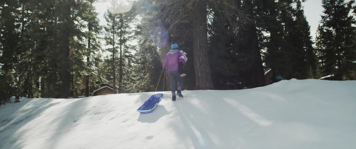 Woman wearing pink coat and holding a child pulls a blue sled up a hill with sun shining brightly through the trees