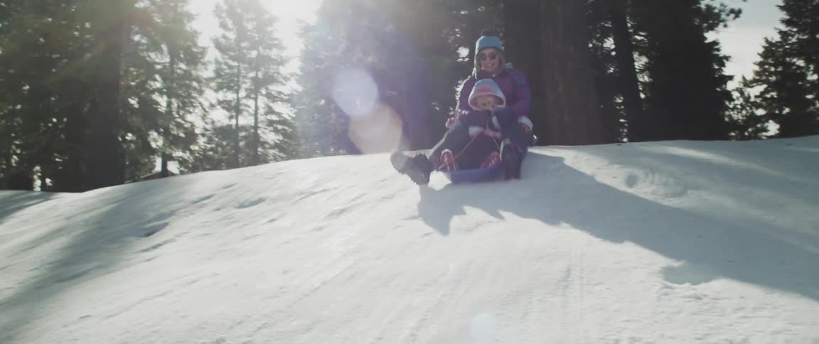 An adult holds a child on a sled while moving down a snowy hill
