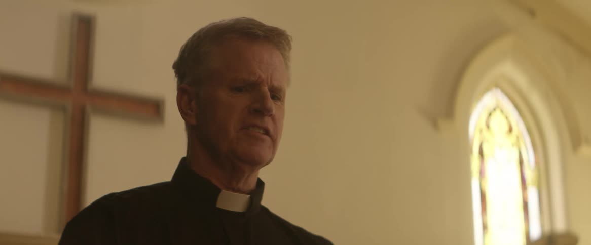 A priest stands in front of a cross indoors talking