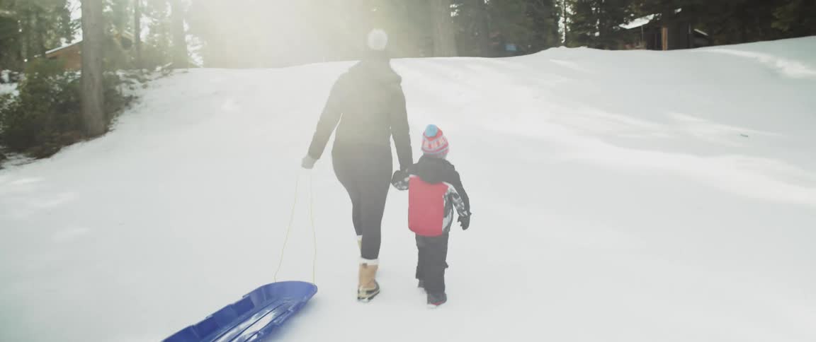 A woman and a child walking on snow while the woman pulls a sled with trees the sky and clouds in the background