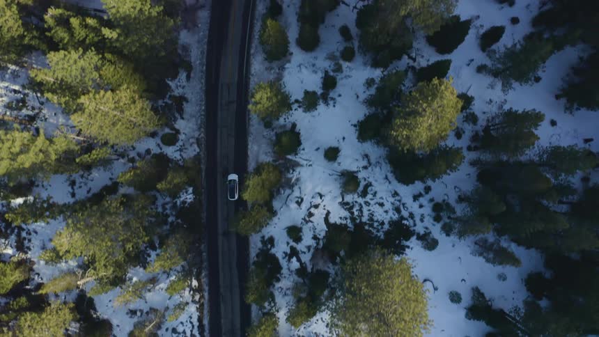A white vehicle drives down a two lane highwaythrough a snow dusted forest