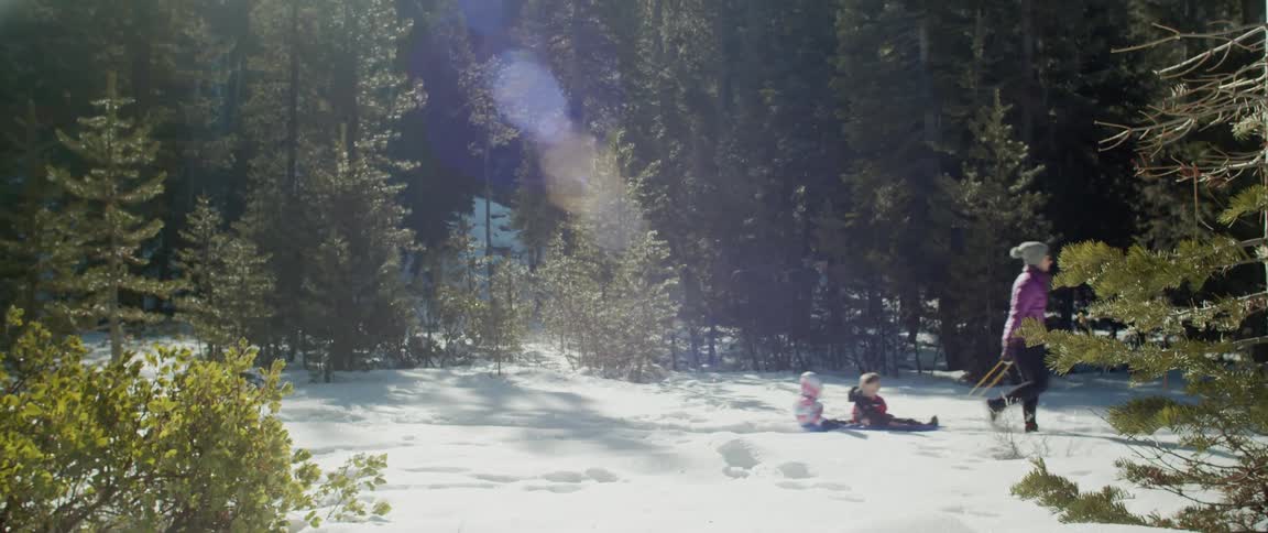 Woman wearing pink coat pulls two children through the snow on a sled with many trees around
