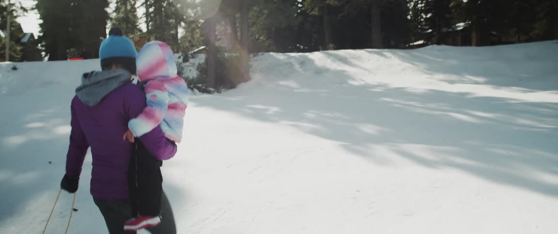A woman holding a child and walking in the snow with trees the sky and clouds in the background