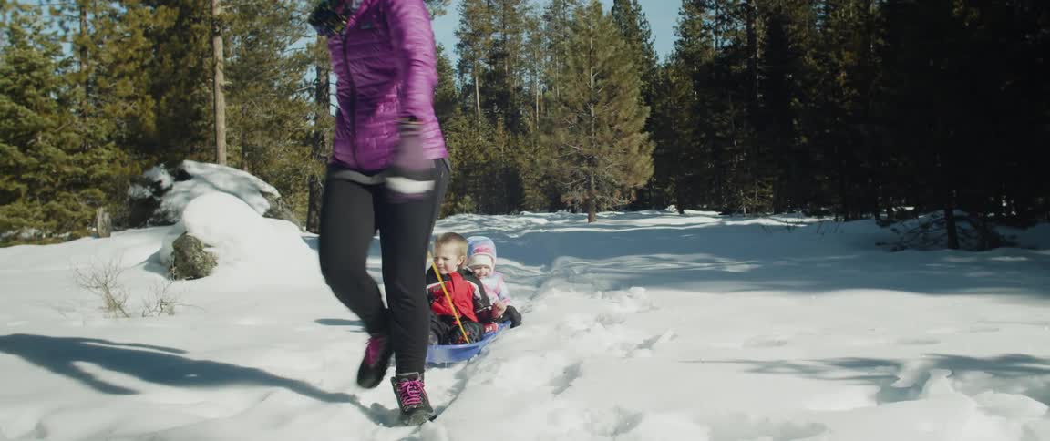 Woman wearing pink coat pulls two young children on blue sled through the snow with many trees around on a clear day