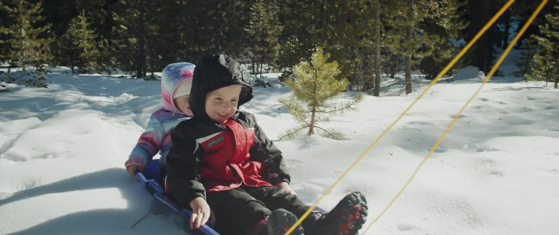 A young boy and a girl sit together on a plastic sled as an adult woman pulls it across a snowy field