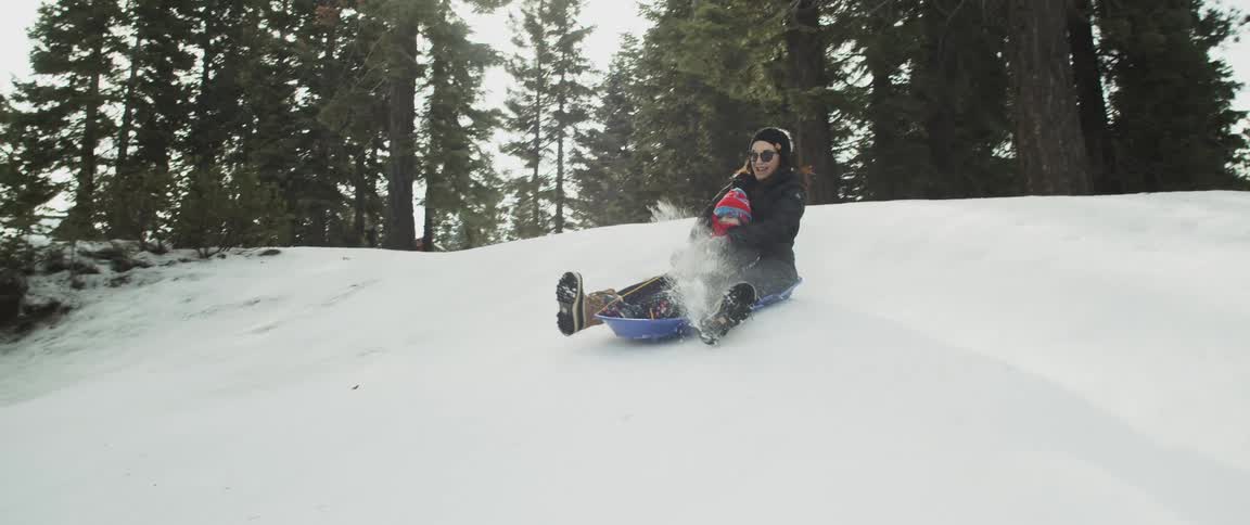 A woman embraces a child as they sled down a snowy hill on a small plastic sled on a winter day