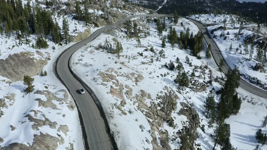 An aerial view of a white car driving on a winding road in a snowy forest