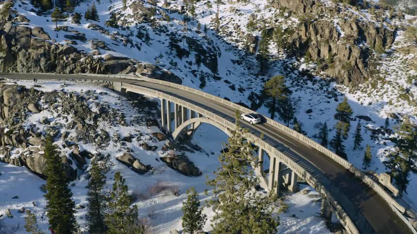 An aerial view of a white car traveling over a bridge in a snowy landscape