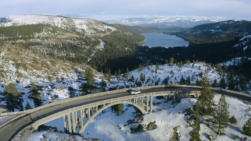 A vehicle drives along a curved bridge that passes by a forest and mountains