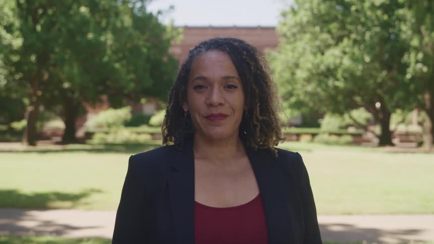 A woman in a suit jacket turns her head and smiles while standing on the lawn of a large campus with full green trees