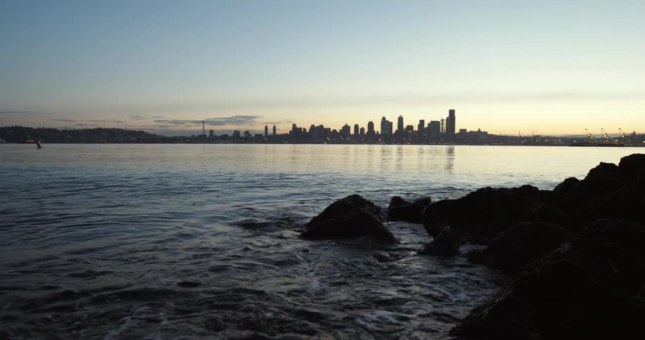 Water crashes against rocks as the sun sets behind a city across the water