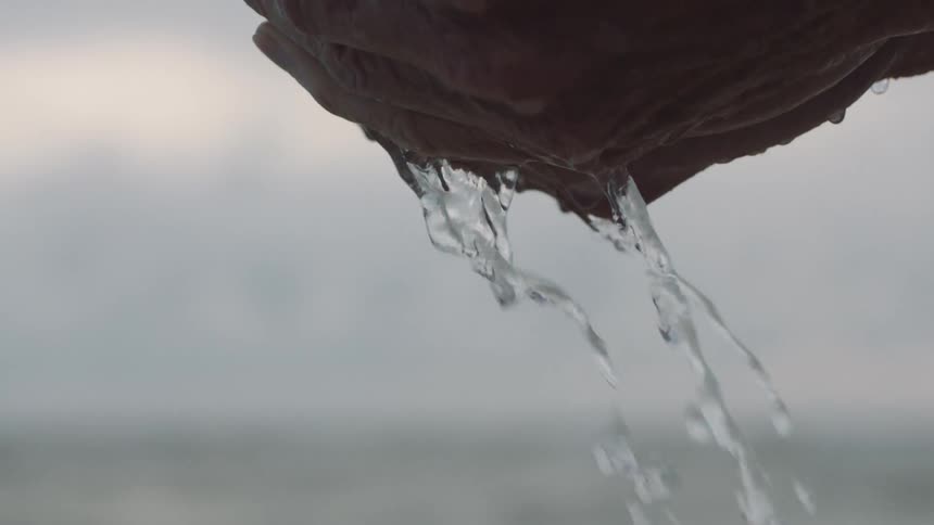 A persons hands holding and dripping water with the ocean the sky and clouds in the background