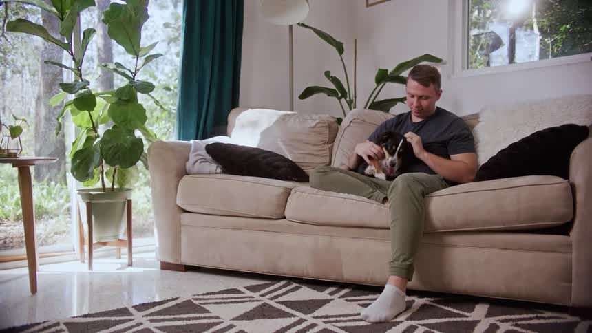 A man pets a dog while sitting on a sofa in the living room of a house during the day