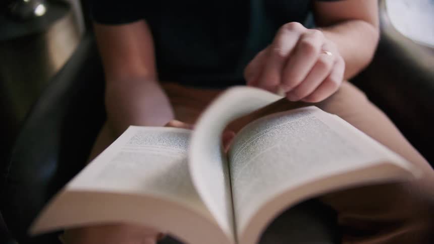 A male is sitting and reading a book while slowly turning the page