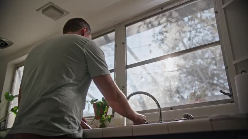 A man turns off water to his sink and scrubs a plate while standing in the kitchen