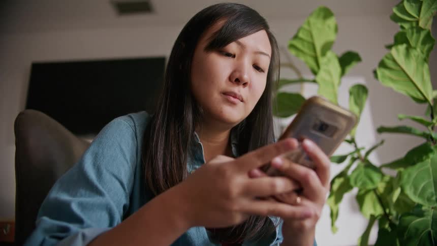 A woman sitting indoors looks down at her smart phone screen while concentrating