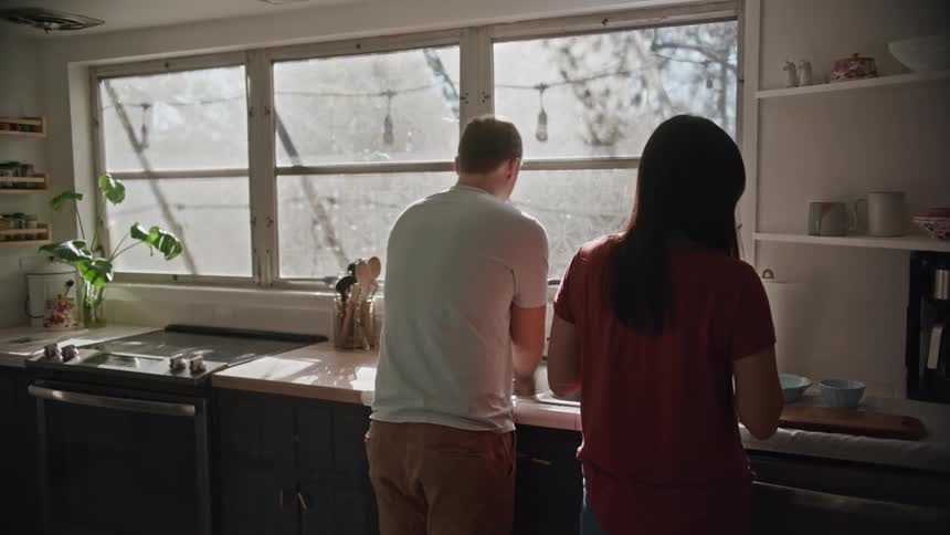 Couple washes dishes together standing in kitchen near window