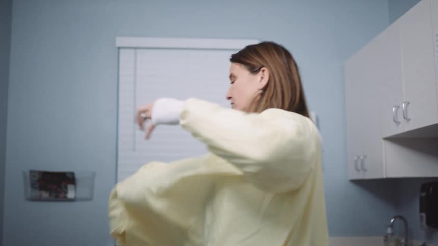 A female doctor in a small doctors office puts on a yellow surgical apron