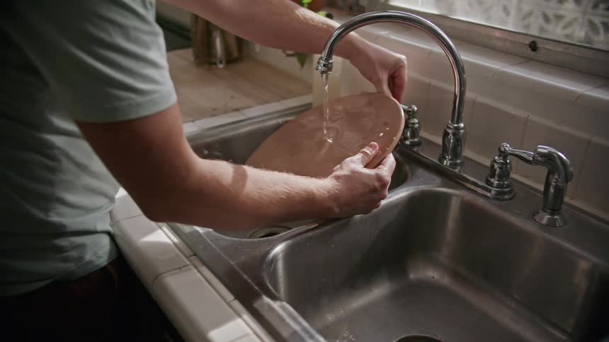 A man rinses a wooden pizza dish under a kitchen faucet before sitting it on the counter to dry