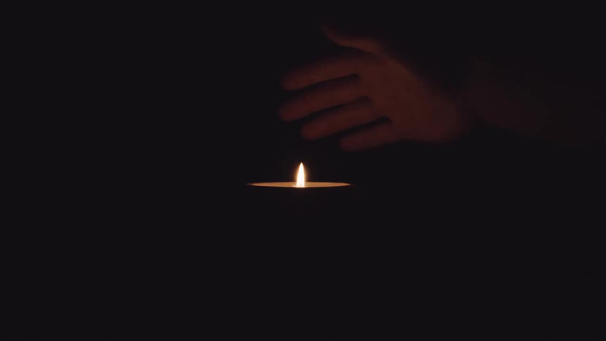 A persons hand over a candle and a flame in the darkness and then outing the fire