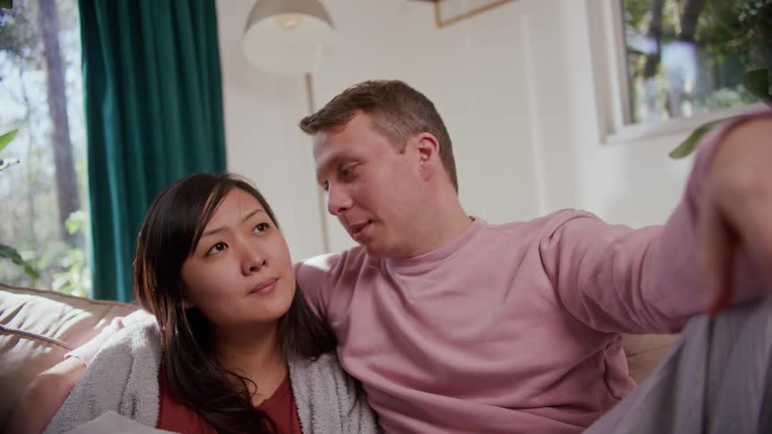 A man and a woman in a room sitting on a couch with plants and a lamp in the background
