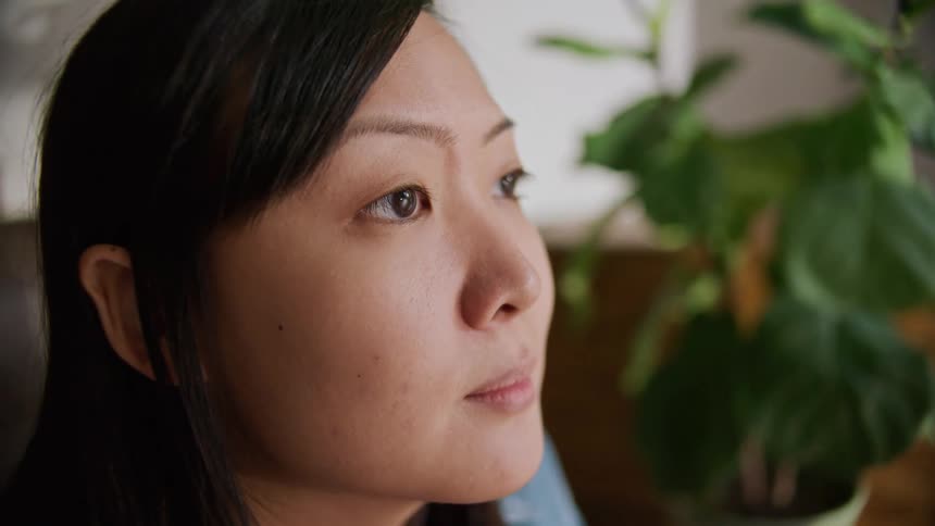 A young asian woman sits inside a house while lost in thought
