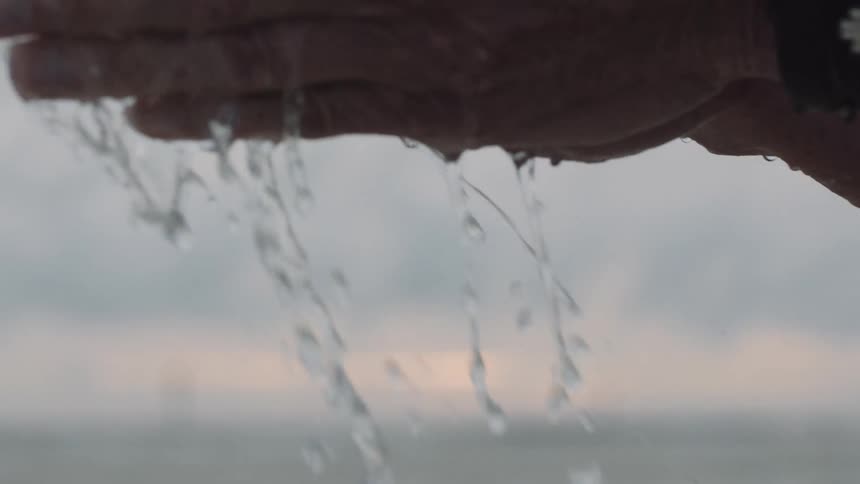 A persons hands holding and dripping water on a beach with the sky and clouds in the background