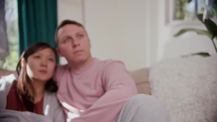A man sits on a couch at home with his arm around his wife and points a remote control in front of him