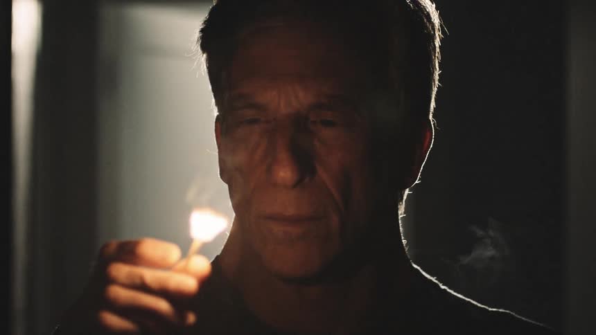 A man standing in the dark is illuminated with light as he lights a match and a door opens