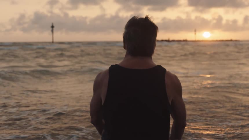 A man stands waist deep in ocean water as the sun hangs low in the sky