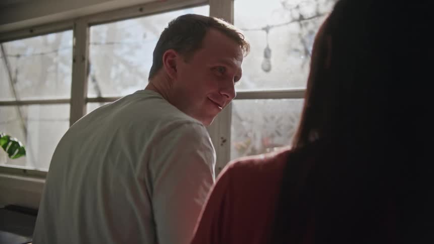 A man stands in front of a sink while washing dishes and smiles at his female companion