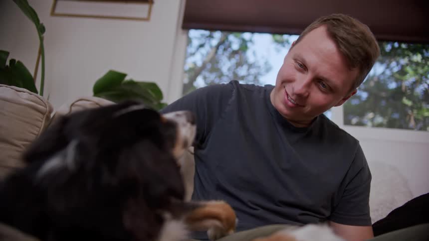 A man pets a dog while sitting on the sofa in the living room of a house
