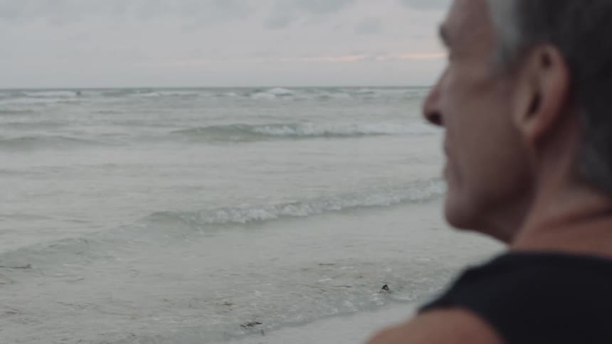 Man wearing black tank top shirt is watching the waves roll in on a cloudy day