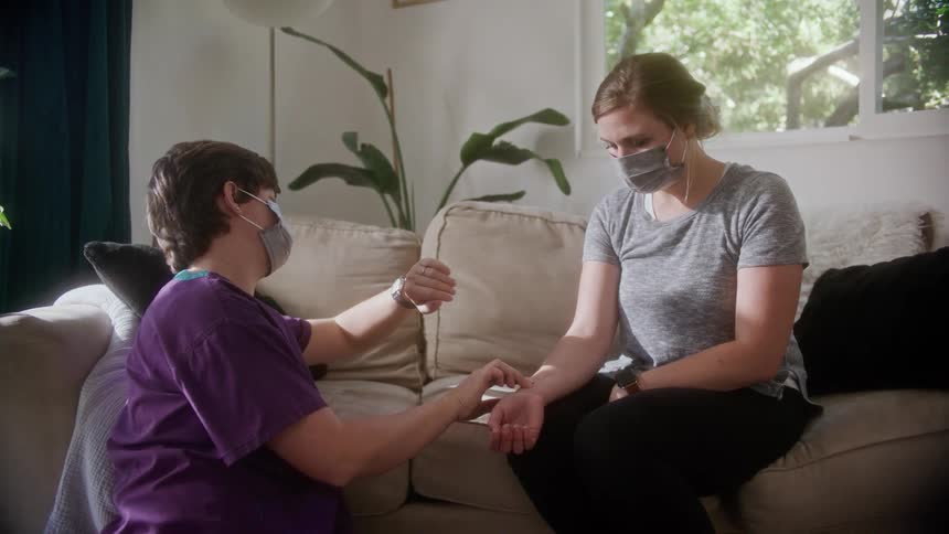 A woman with a mask checking the pulse of another woman sitting on a couch in a room with plants and windows in the background