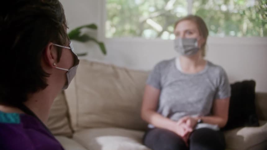 A home health nurse sits while talking to a woman sitting on the couch across from her