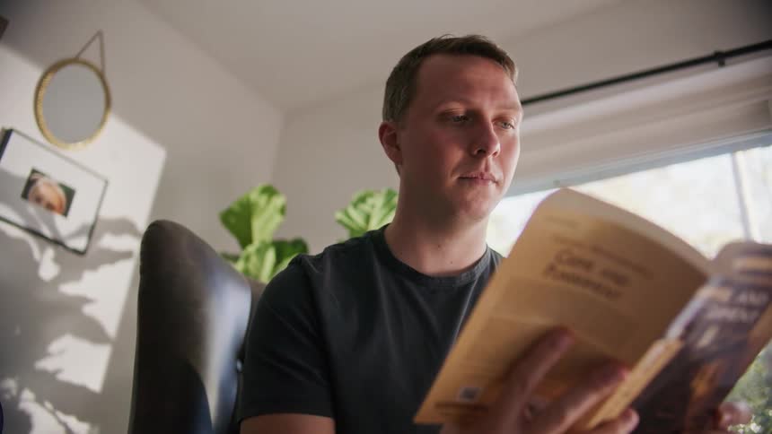 A man sits in a leather chair at home and reads a book near a large window