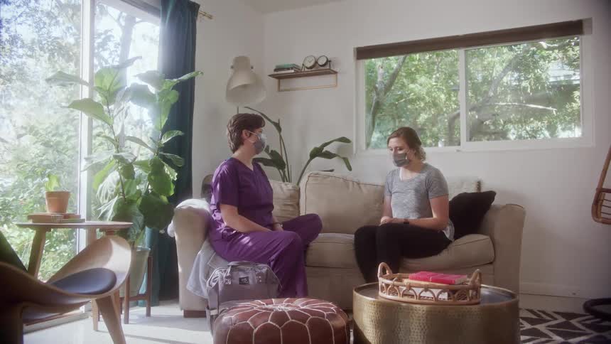 Two women sit together on a home couch talking and wearing face masks