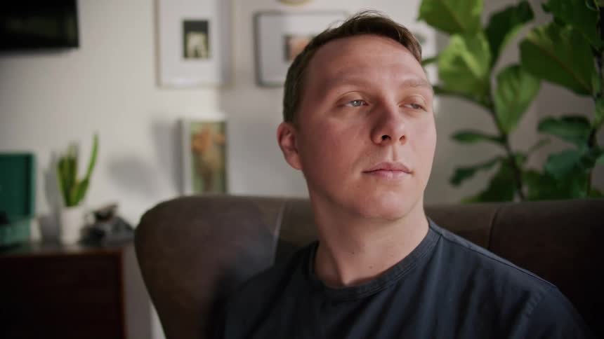 A man in a room sitting on a couch with plants and a wall in the background