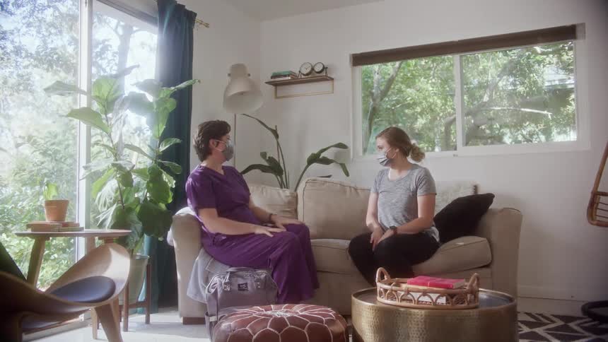 A woman at home sits down on a couch and then a nurse joins her and they talk