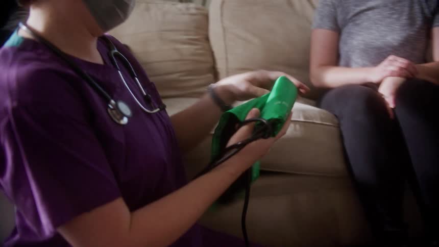 A nurse with a stethoscope and a mask checking the blood pressure of a woman sitting on a couch