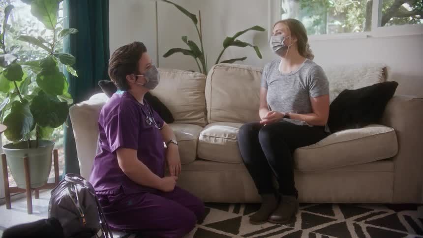 A young woman sitting on her couch as home points to her stomach as she talks to a nurse