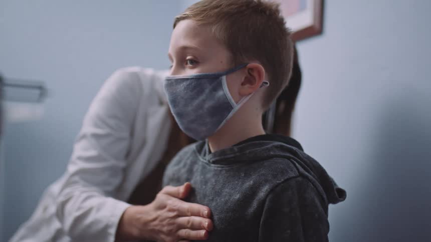 A doctor with a mask in a clinic using a stethoscope on a boy