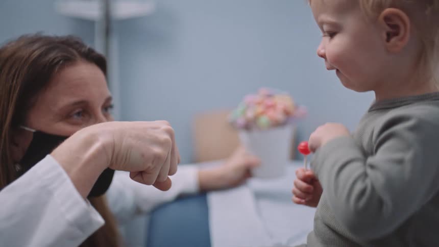 A doctor with a mask giving a fist bump to a little girl with a lollipop in a clinic