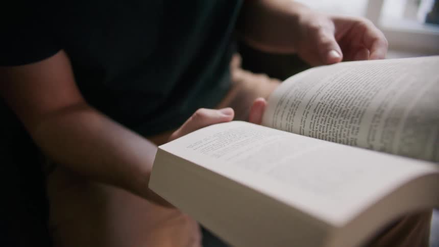 A man turns the page of the book he is reading indoors during the day