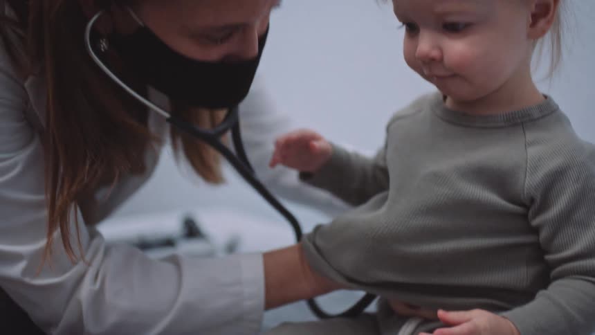 A female doctor shows the end of a stethoscope to a small girl sitting on an examination table before using it to listen to the girls chest