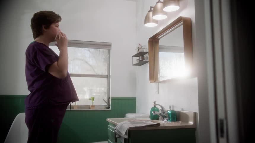 A nurse looks in a bathroom mirror while putting on a gray face mask