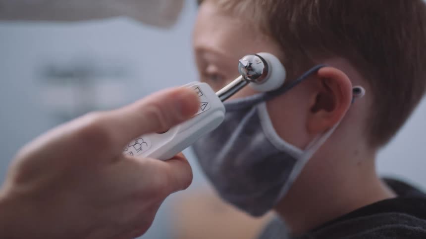 A doctor swipes a thermometer across a boys forehead as he sits on the exam table wearing a mask
