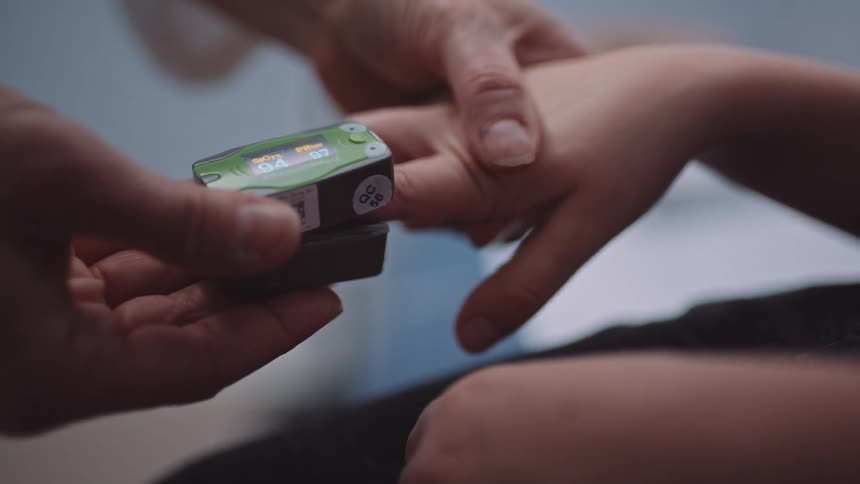 A person in a room using a device on a childs finger with a wall in the background