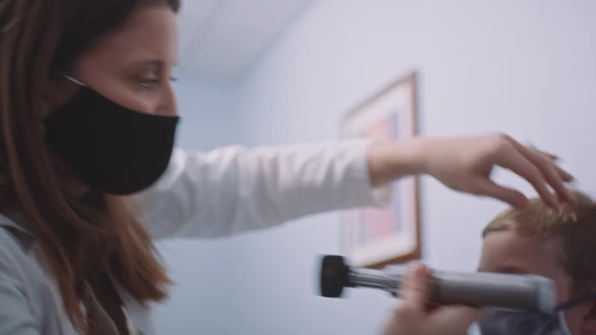A female doctor in a medical mask uses an otoscope to examine the ears of a masked boy sitting in her office